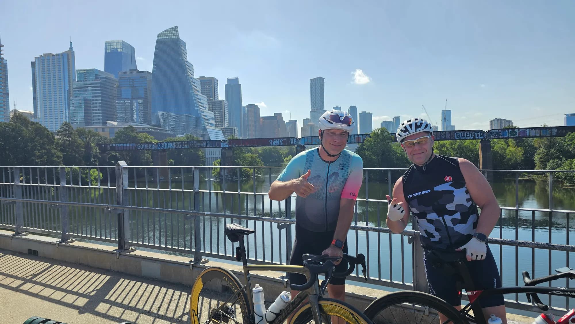 Two riders smiling with their bikes on a bridge over the water, the downtown Austin skyline behind them
