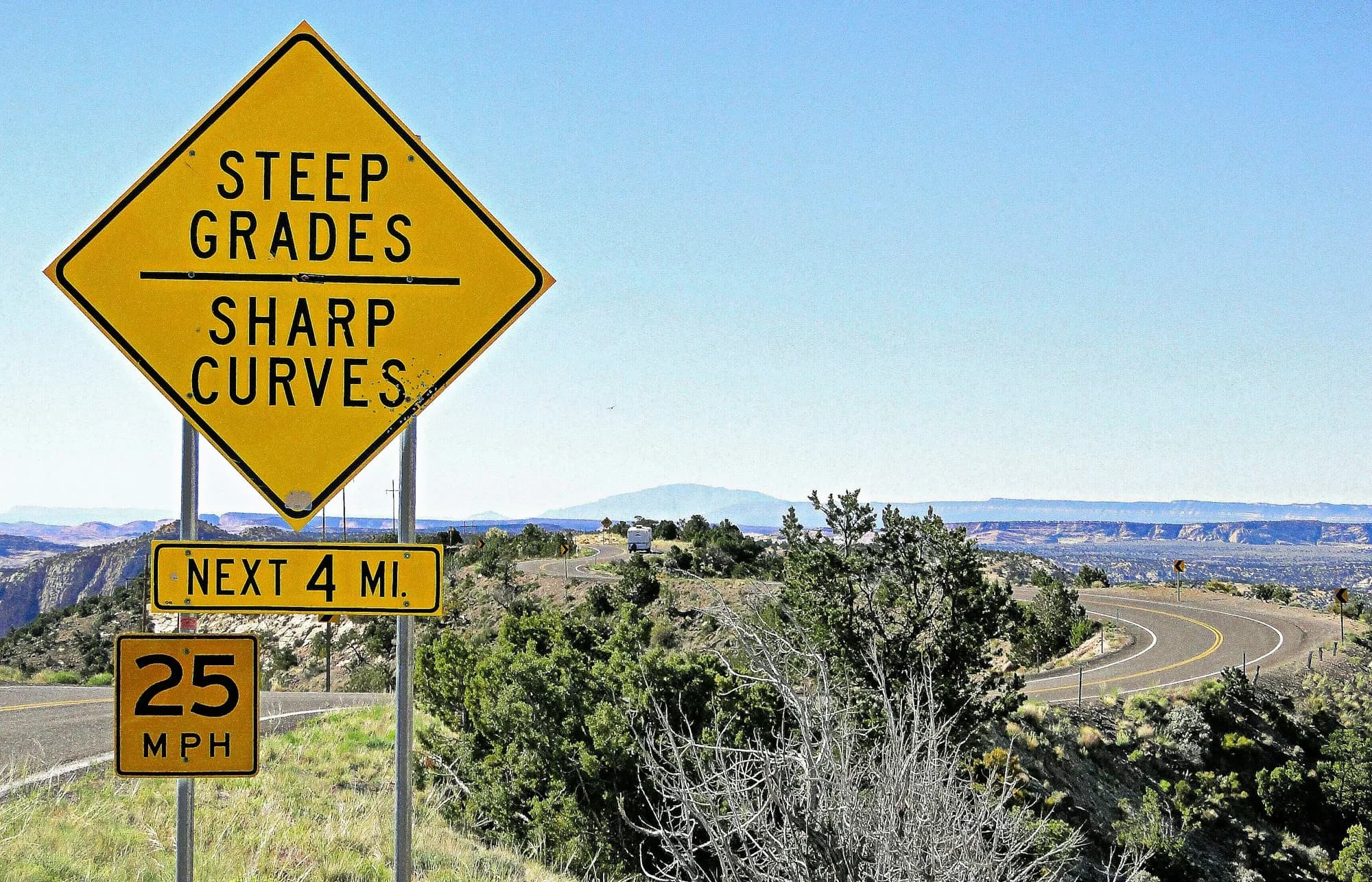 A yellow road sign reading Steep Grades, Sharp Curves, Next 4 Mi beside a winding mountain road
