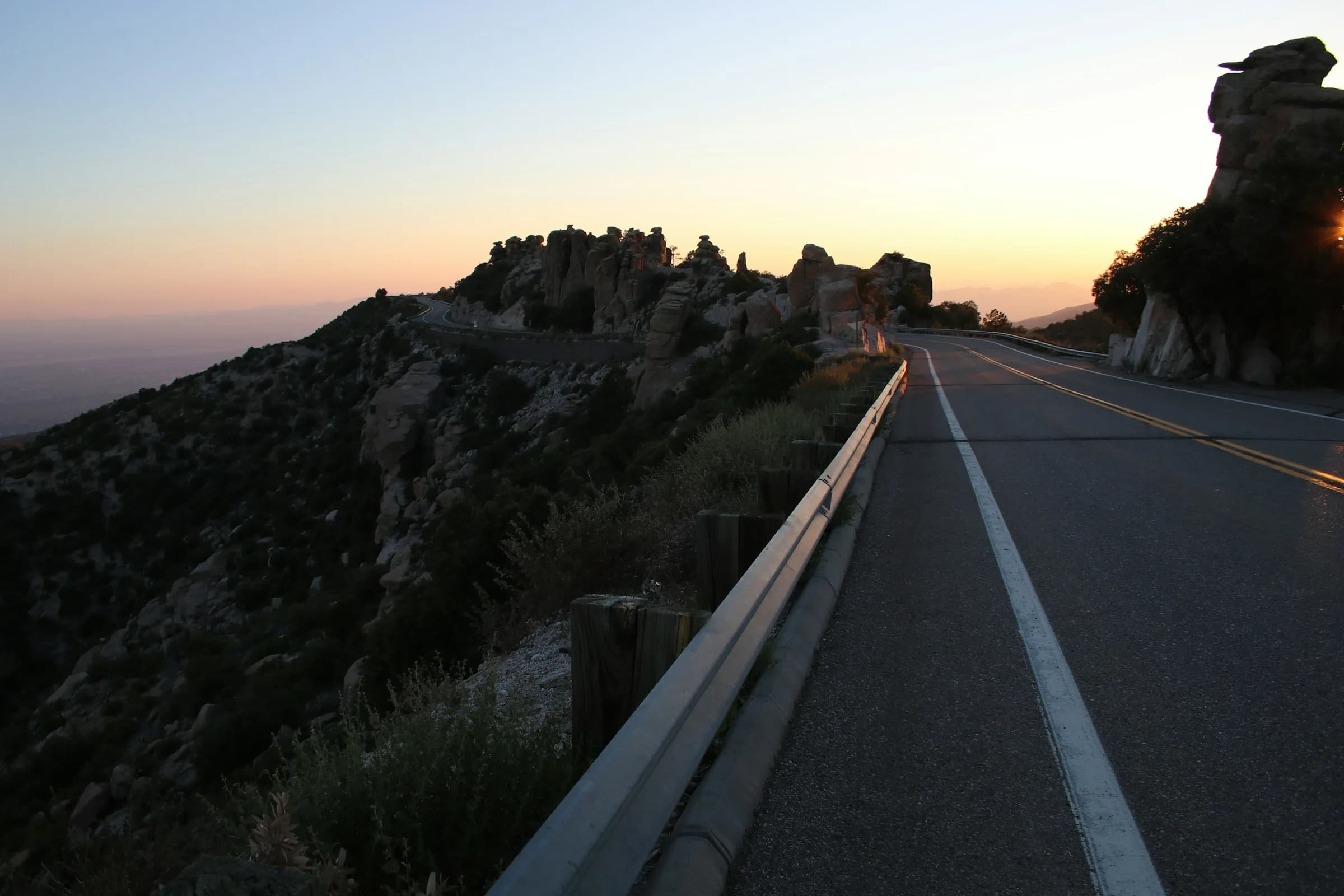 A paved mountain road with a guardrail running toward rock formations at dusk