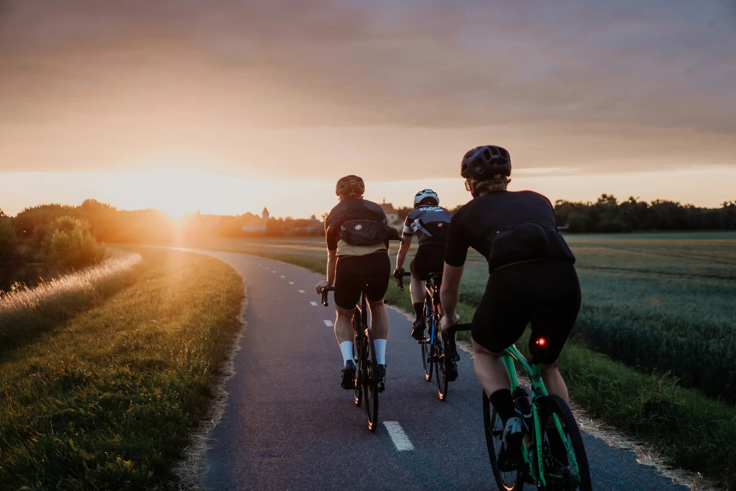 Three road cyclists riding away along a paved path toward the setting sun