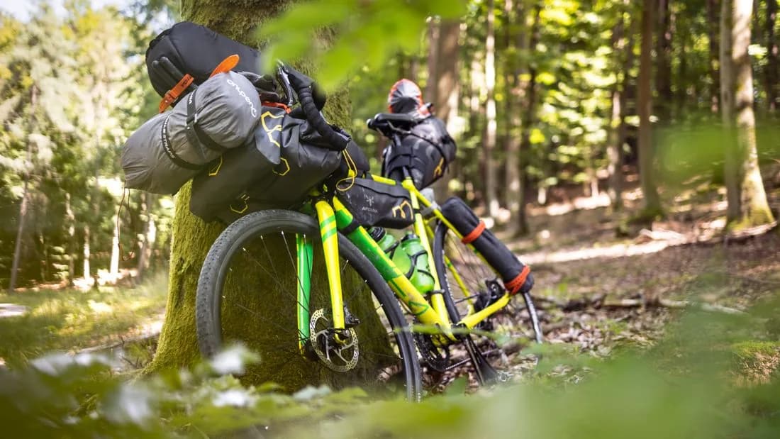 A bicycle loaded with bikepacking bags, a sleeping bag strapped across the handlebars, leaning against a tree in a forest